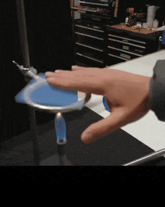 Ръка бързо почуква въртящ се син диск, монтиран на метален прът, което го кара да се върти по-бързо. На заден план показва обстановка на работилница с инструменти и оборудване.