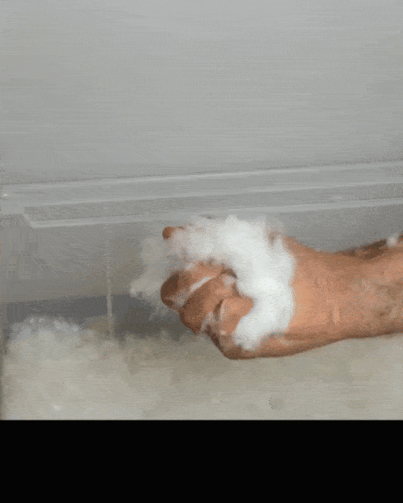 Човек изстисква бяло пухкаво вещество от ръката си под вентилационен отвор, освобождавайки мек на вид, подобен на памук материал. Веществото пада бавно и се разпръсква във въздуха.