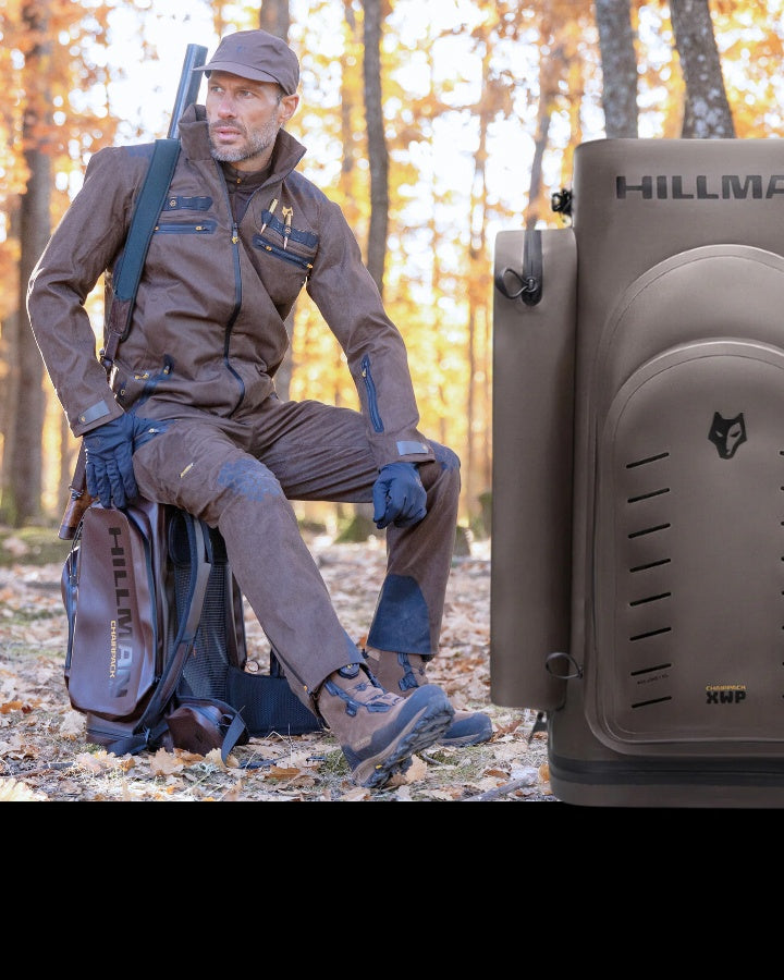 Човек, облечен в кафява екипировка за открито и шапка, стои в горска среда през есента. Подпрени са на раница, на която се вижда марката "Hillman". Златни листа покриват земята, а дървета с оранжева зеленина са на заден план.